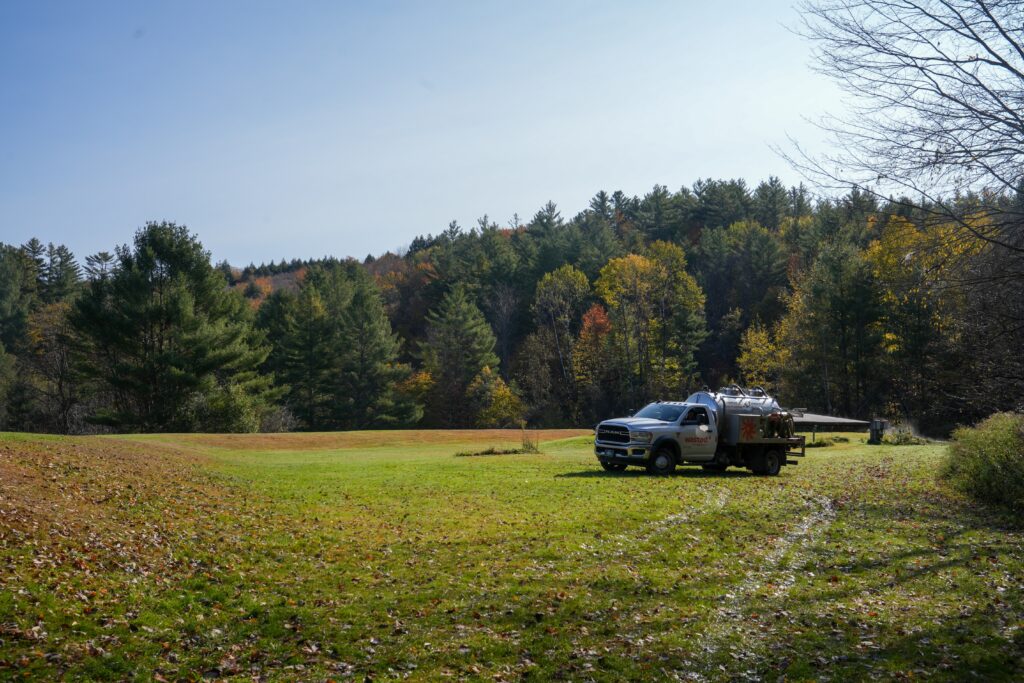 Wasted* service truck parked in a grassy field near a Vermont forest, with sanitation equipment onboard—showcasing offgrid portable toilet support in rural New England during fall.